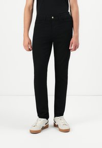 Μαύρα slim-fit τζιν με λεία υφή, πέντε τσέπες και κλείσιμο με κουμπί μπροστά. Συνδυάζονται με λευκά αθλητικά παπούτσια και μαύρο πουκάμισο.