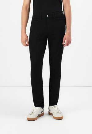 Black slim-fit jeans with a smooth texture, five pockets, and a front button closure. Paired with white sneakers and a black shirt.