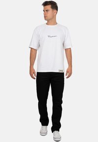 T-shirt bianca in cotone con logo a scritta nera, abbinata a jeans neri e sneakers bianche. La t-shirt ha maniche raglan e una vestibilità comoda.