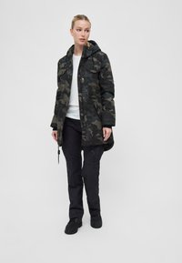 Kamikaze jakas tumši zaļā un pelēkā krāsā, ar kapuci, priekšējām kabatām un auklām ap apakšmalu. Kombinēta ar melniem biksēm un apaviem.