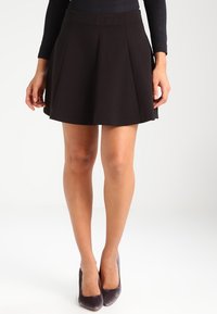 Falda plisada negra de tela, con una cintura ajustada y forma acampanada, combinada con tacones altos de ante gris.