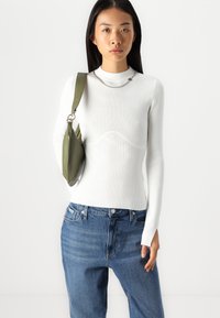 Balts ribots garām piedurknēm džemperis ar augstu kakla izgriezumu, kuram ir viļņota tekstūra ķermeņa daļā, saskaņots ar ziliem džinsu biksēm un zaļu plecu somu.