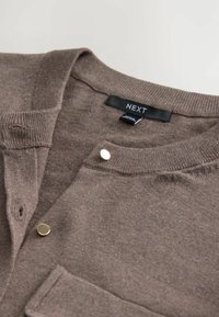 Hnědý pletený cardigan s kulatým výstřihem, se zlatým zapínáním na knoflík a malou kapsičkou na prsou. Látka vypadá měkce a má strukturovaný povrch.