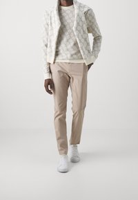 Cremefarbener Kapuzenpullover mit Reißverschluss über einem cremefarbenen Pullover, kombiniert mit hellbraunen Hosen und weißen Turnschuhen. Glatte Texturen und modernes Design.