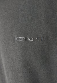 Camiseta de algodón gris con textura acanalada, que presenta el logo bordado en blanco "carhartt" en el frente. Diseño simple, estilo casual.