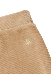 Tan corduroy stof met verticale ribbels. Bevat een klein geborduurd logo. Zachte textuur en matte afwerking. Close-up van casual kledingdetail.