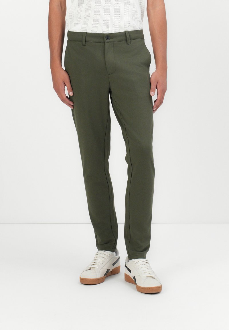 Pantalones ajustados verde oliva con un corte slim, cierre de un solo botón y bolsillos frontales; combinados con zapatillas blancas y una blusa de punto texturizada.