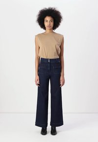 Top sem mangas em tom bronze, combinado com calças de ganga largas e escuras, apresentando bolsos frontais e dois botões. A modelo está de pé contra um fundo simples.