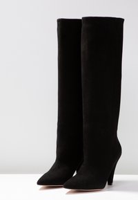 Une paire de bottes noires en daim hautes avec des pointes et des talons hauts sur une surface blanche contre un fond gris.