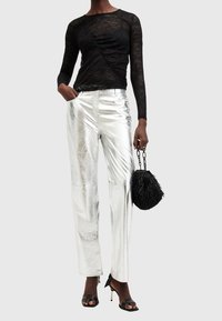 Femme portant un haut à manches longues en dentelle noire, pantalon argenté métallique, sandales à talons noirs, tenant un sac à main noir texturé avec des anses en chaîne.