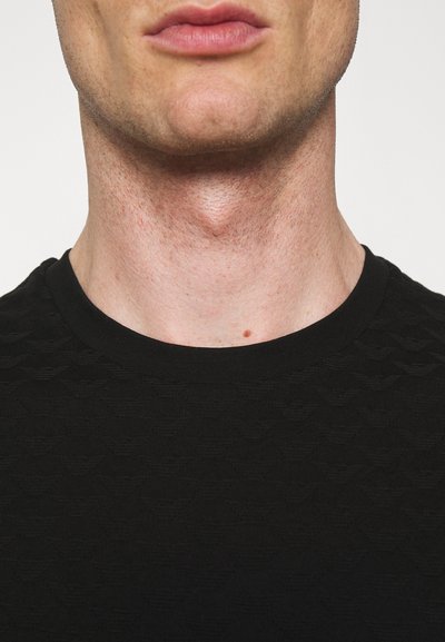 Camiseta negra texturizada con cuello redondo, que presenta un patrón en relieve. Primer plano del cuello y área superior del pecho. Tejido suave.