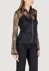 Blusa di pizzo nero con un design floreale trasparente, maniche lunghe e colletto con bottoni. Il tessuto ha un aspetto testurizzato; silhouette aderente.