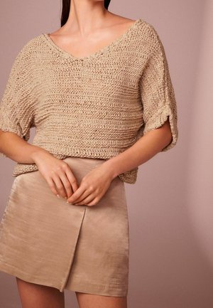 Femme portant un pull en tricot beige à manches courtes amples et une mini-jupe en satin marron clair avec une fente à l'avant, debout devant un fond neutre.