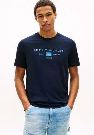 Giovane uomo che indossa una t-shirt Tommy Hilfiger blu navy con logo blu e jeans azzurri, in piedi con le mani nelle tasche.