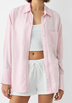 Kobieta w dużej koszuli na guziki w różowo-białe paski, na białym krótkim topie i białych szortach ze ściągaczem.
