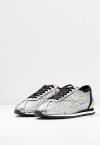 Sneakers in rete argentato con accenti neri, lacci piatti e motivo testurizzato, caratterizzati da un profilo basso e una suola nera slim e resistente.