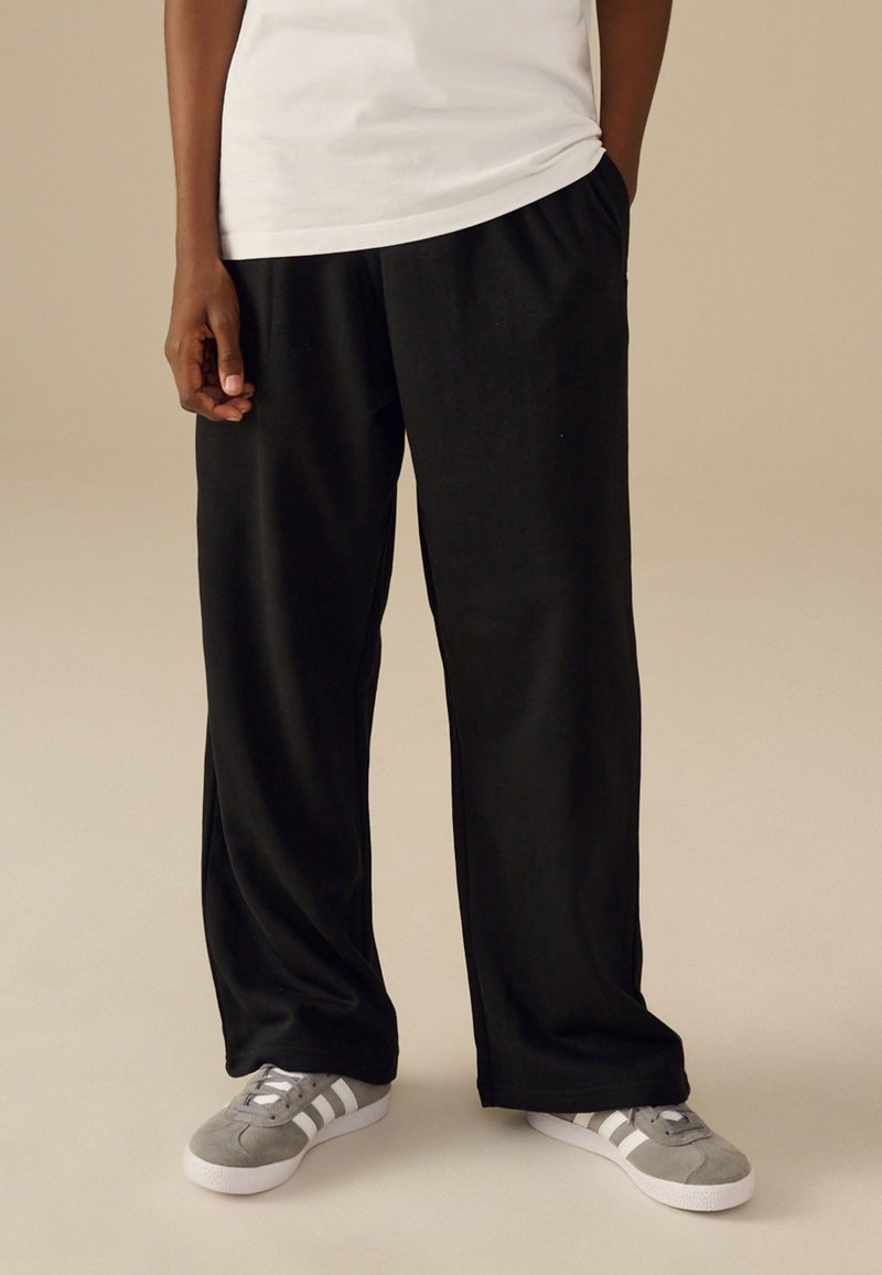 Persona che indossa pantaloni larghi neri, maglietta bianca e sneakers grigie con strisce bianche, in piedi su pavimento beige con sfondo beige.