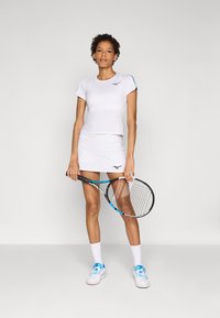 Bela atletska majica in krilo z modrim akcentom, s prilegajočim dizajnom. Model drži tenis lopar in nosi bele čevlje ter nogavice.