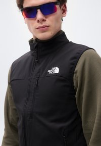 Νέος άνδρας που φοράει αντανακλαστικά αθλητικά γυαλιά ηλίου, μαύρο γιλέκο με φερμουάρ και το λογότυπο "The North Face" και υφασμένο μακρυμάνικο πουκάμισο ελιάς.