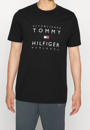 Schwarzes Baumwoll-T-Shirt mit Rundhalsausschnitt, versehen mit weißem Text "ESTABLISHED TOMMY HILFIGER" und einem rechteckigen Logo in Rot, Weiß und Blau.
