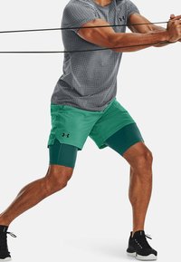 Zelené atletické šortky s vrstveným designem a délkou ke kolenům, spárované s šedým tričkem s krátkým rukávem a texturou, nošené během cvičení.