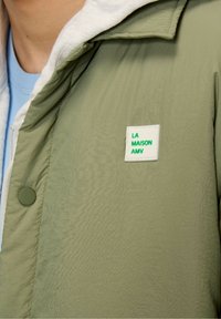 Lys grøn jakke med en struktureret overflade, der har en hvid foring og et rektangulært mærke, der står "LA MAISON AMV" i grøn. Knaplukning.