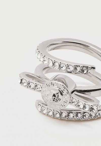 Σωρευμένα ασημένια δαχτυλίδια με διάφανα κρύσταλλα, ένα δαχτυλίδι με μοτίβο ελληνικού κλειδιού και κεντρικό έμβλημα κεφαλής λιονταριού.