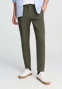 Oliwkowe spodnie o slim fit, wykonane z bawełny. Uzupełnione białymi trampkami z beżową podeszwą oraz lekką, niezapiętą niebieską koszulą.