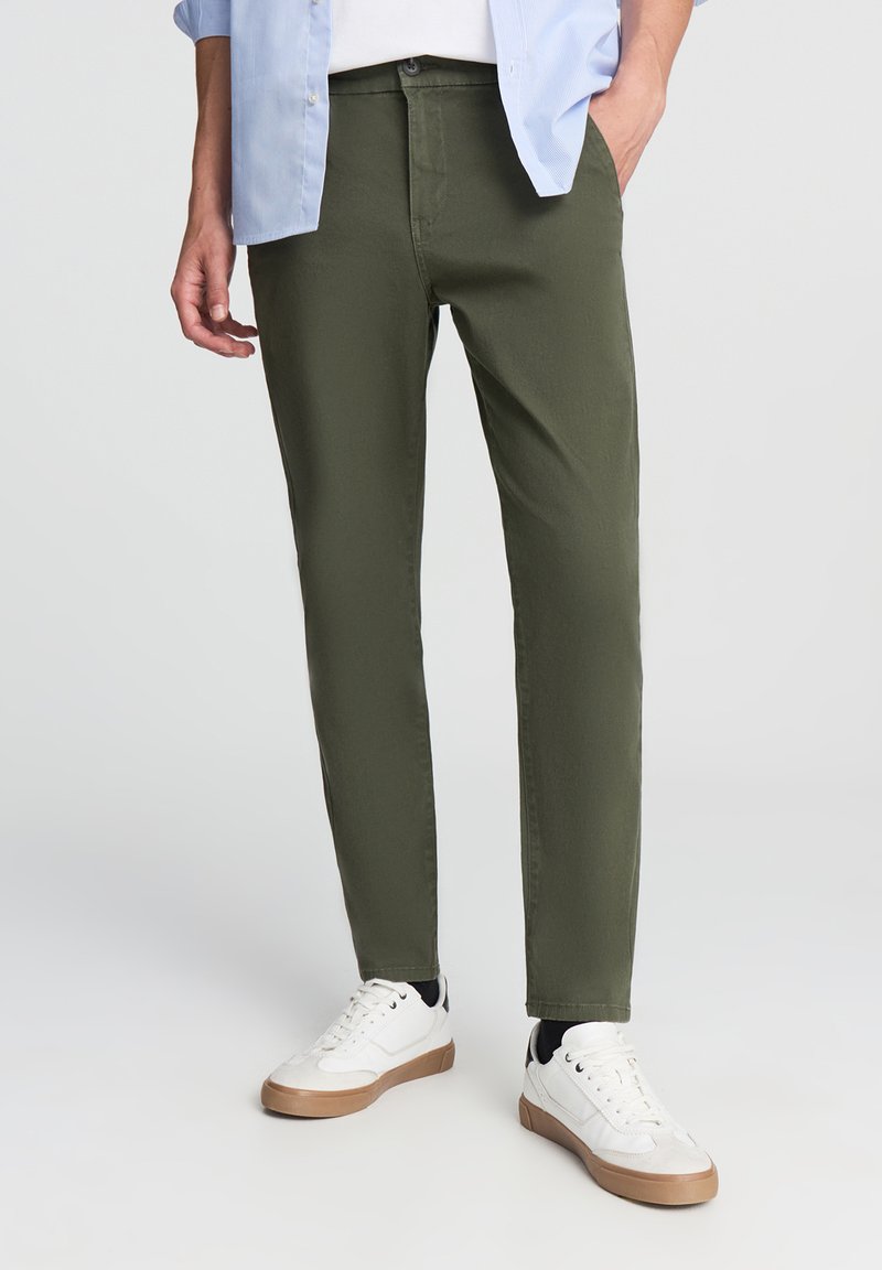 Oliwkowe spodnie o slim fit, wykonane z bawełny. Uzupełnione białymi trampkami z beżową podeszwą oraz lekką, niezapiętą niebieską koszulą.