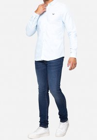 Camisa de botones azul claro con mangas largas, combinada con jeans ajustados azul oscuro y zapatillas blancas. Incluye un pequeño logo en el pecho.