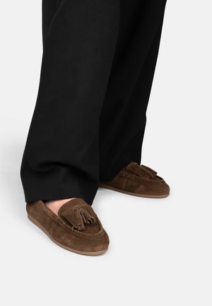 Gambe che indossano pantaloni neri larghi e mocassini marroni in camoscio con nappine e suola in gomma su un pavimento bianco.