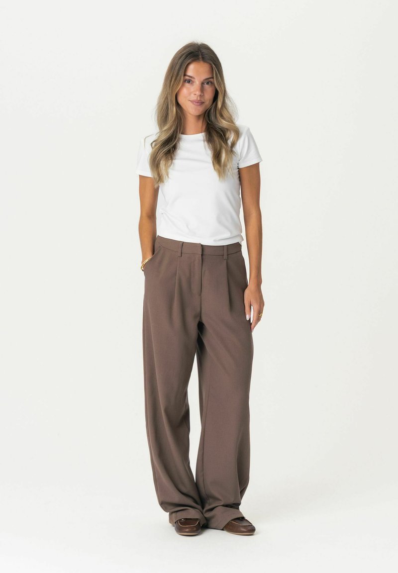 T-shirt bianco a maniche corte abbinato a pantaloni larghi, a vita alta, di colore marrone. I pantaloni presentano una parte anteriore plissettata e una texture liscia.