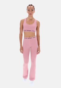 Jasnoróżowy zestaw sportowego stanika i dzwonowatych legginsów z kontrastującą czarną opaską z logo. Gładki, elastyczny materiał i dopasowany krój. Białe buty.