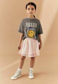 Camiseta gris de algodón con el texto "NIRVANA" y un gráfico de carita sonriente, combinada con una falda iridiscente en tonos pasteles y zapatillas plateadas de caña alta.