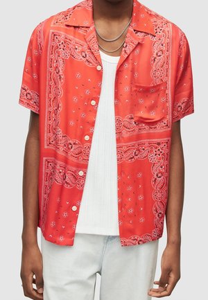 Mand iført rød kortærmet skjorte med hvid paisley-mønster over hvid ribbet tanktop og lyseblå jeans.