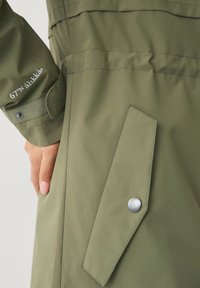 Cappotto verde oliva con una texture liscia, dettagli con bottoni a pressione e branding ricamato sulla manica. Design su misura con un taglio alla moda.