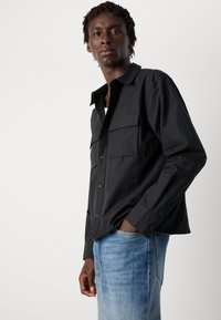 Svart langermet skjorte med to frontlommer, knappestop, og avslappet passform, kombinert med lyseblå jeans som viser en glatt tekstur.