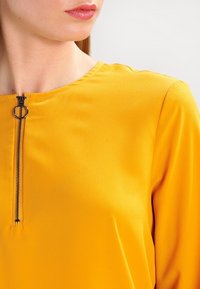 Žlutý top s hladkou texturou, kulatým výstřihem a předním zipem s malým kovovým kroužkem pro snadné zapínání.