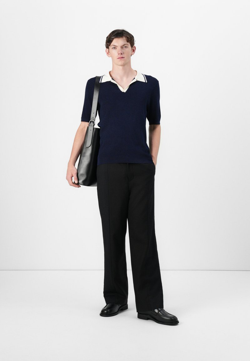 Navyblaues Polohemd mit kurzen Ärmeln und weißem Kragen, kombiniert mit maßgeschneiderten schwarzen Hosen und schwarzen Schuhen. Eine schwarze Umhängetasche hängt über der Schulter.