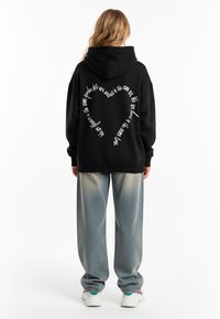 Felpa nera oversize con cappuccio, caratterizzata da un testo bianco disposto a forma di cuore sulla schiena. Abbinata a jeans baggy azzurri e sneakers.