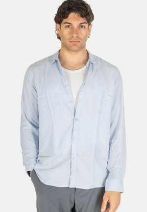Giovane uomo con capelli ricci scuri che indossa una camicia azzurro chiaro sbottonata sopra una canottiera bianca, in piedi davanti a uno sfondo uniforme.