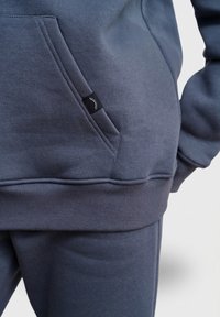 Tmavě šedá mikina s kapsou vpředu, žebrovanými manžetami a pasem. Na kapse je malá černá etiketa s bílým logem.