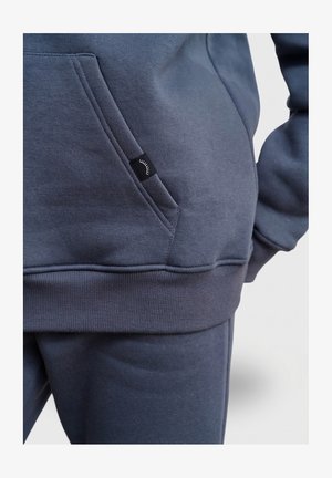 Tmavě šedá mikina s kapsou vpředu, žebrovanými manžetami a pasem. Na kapse je malá černá etiketa s bílým logem.