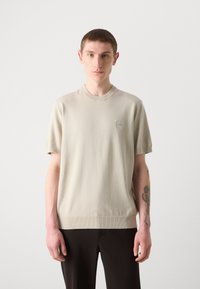 Beige rövid ujjú pulóver, kerek nyakkivágással, texturált anyagból készült. Kicsi logós címkével rendelkezik a mellkason és bordázott mandzsettákkal.