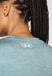 Türkiis spordipoliisil, millel on tekstuur, tagaküljel kaeluse lähedal valge Under Armouri logo.