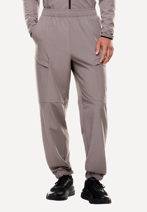 Uomo che indossa pantaloni cargo jogger color taupe con vita elastica e polsini, abbinati a scarpe da ginnastica nere, mani rilassate ai lati.