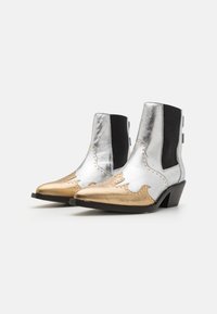 Botas de tobillo de cuero metálico plateado con lados elásticos negros y puntera dorada en forma afilada, que presentan un contraste de costuras en forma de ola y tachuelas.