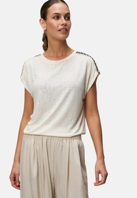 Lysbeige t-shirt med løs pasform, korte ærmer og sorte blonder detaljer på skuldrene. Sat sammen med bløde, cremede bukser.