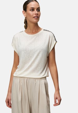 T-shirt beige clair avec une coupe décontractée, des manches courtes et des accents en dentelle noire sur les épaules. Associé à un pantalon doux couleur crème.