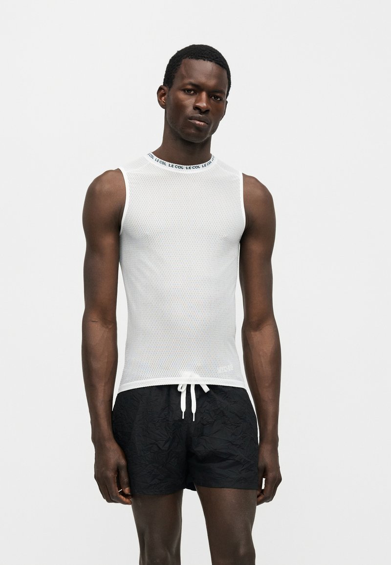 Modèle masculin portant un haut en mesh blanc sans manches avec col marqué et un short noir à cordon, debout devant un fond clair uni.